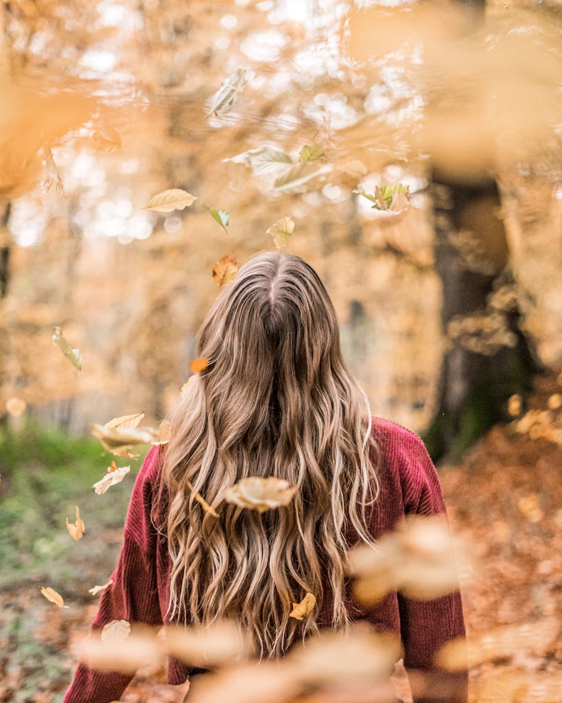 A woman stands among falling autumn leaves in a forest, capturing the essence of fall.