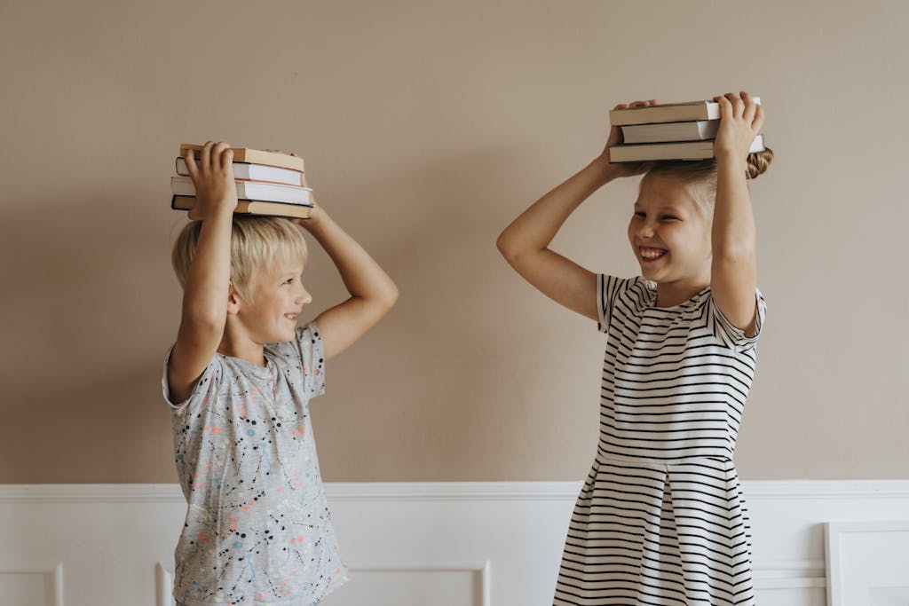 Two smiling children balancing books on their heads indoors, symbolizing fun in learning.